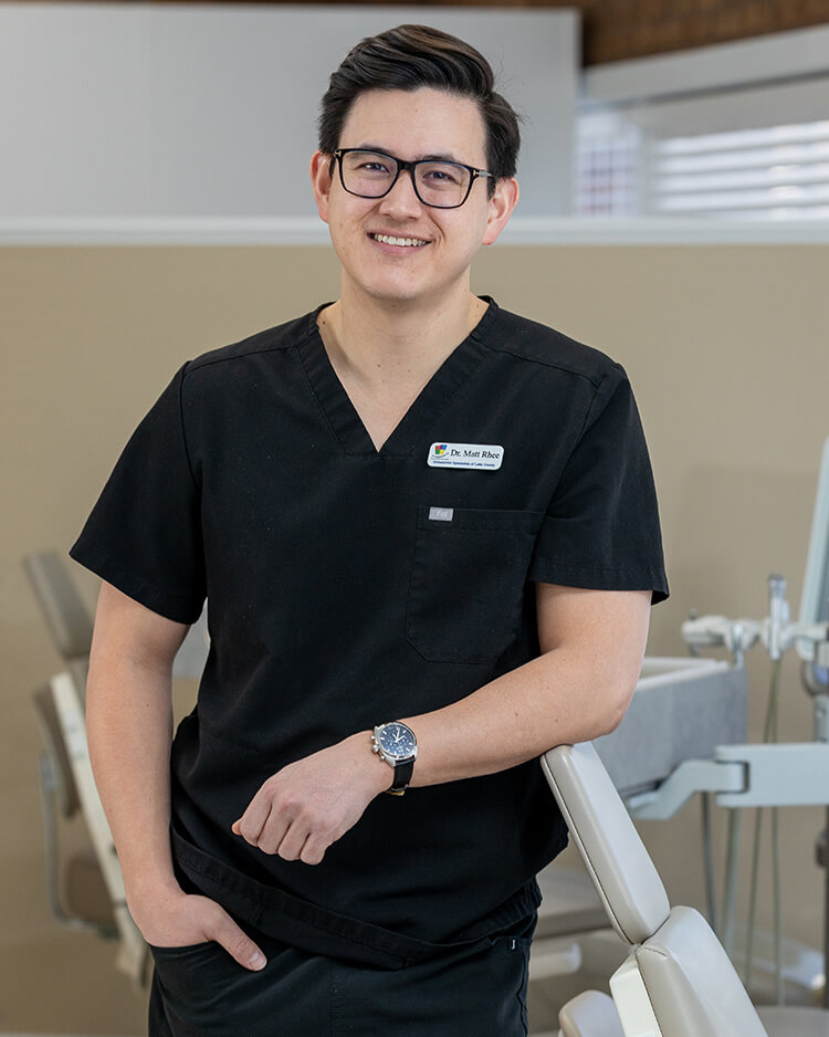 Dr. Rhee, smiling in black scrubs and glasses beside a dental chair