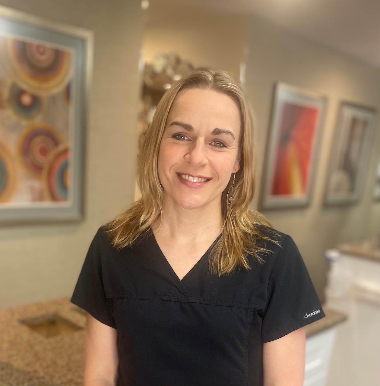 Smiling woman in black scrubs at an office front desk with framed art